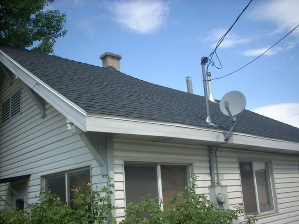 Roof of a house with a chimney, satellite dish, and utility connections under a blue sky.