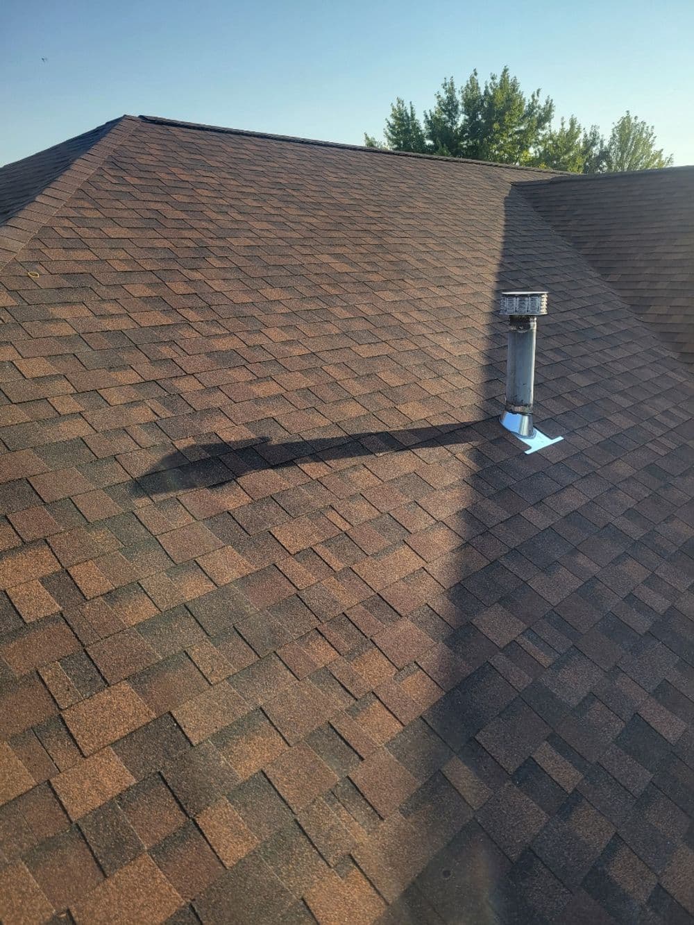 Roof with brown shingles and metal chimney casting a long shadow under a clear blue sky.